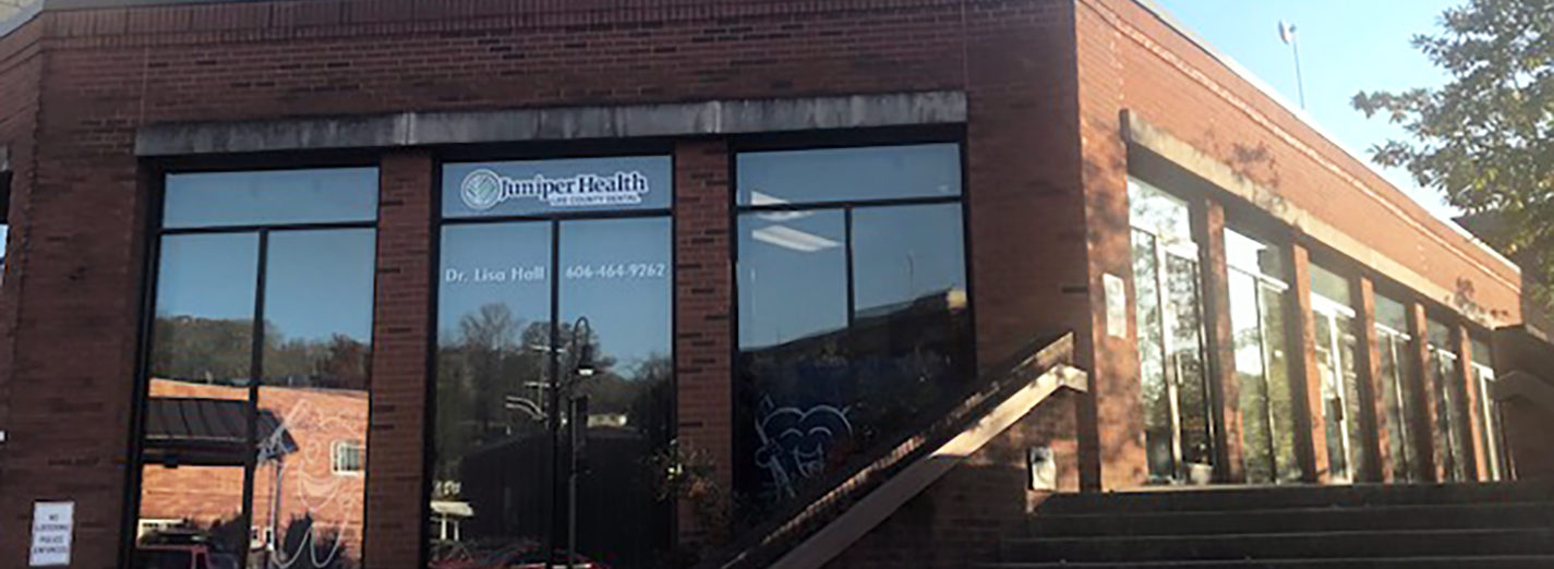 Color photo of the Juniper Health Lee Dental clinic building exterior. Red brick, one-story, with large windows, concrete steps and hand rail. Blue, cloudless sky above, sunny, with reflection of trees in windows. Dark shadows.