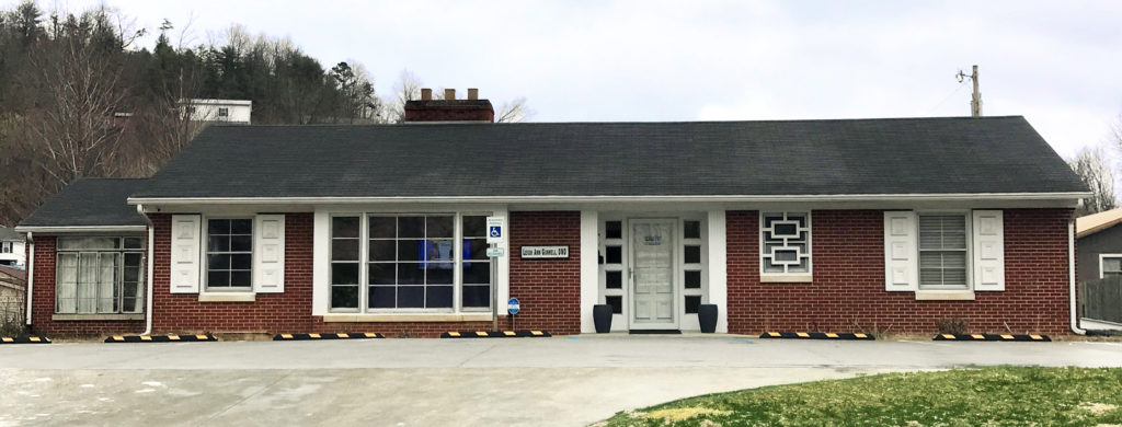 Color photo of the Juniper Health Morgan County Dental Clinic building. Red brick, one-story, black shingled roof. White-trimmed windows and covered doorway. Sky and trees in the background, concrete parking lot in the front.