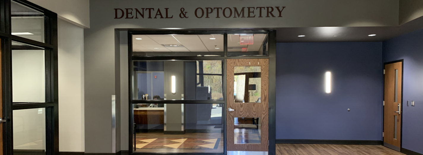 Color photo of interior of Juniper Health Breathitt County clinic, dental and optometry side. Large, glass windows, blue and gray walls, wood floors. Doorway with Dental & Optometry in large, gray letters, above.