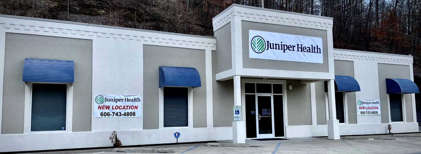 A color photo of the Juniper Health Morgan County Medical Clinic building. Tan and off-white stucco with blue awnings above the dark windows. Covered from door area. Vinyl signs with Juniper Health logo hanging on the front of the building. Trees in the background.
